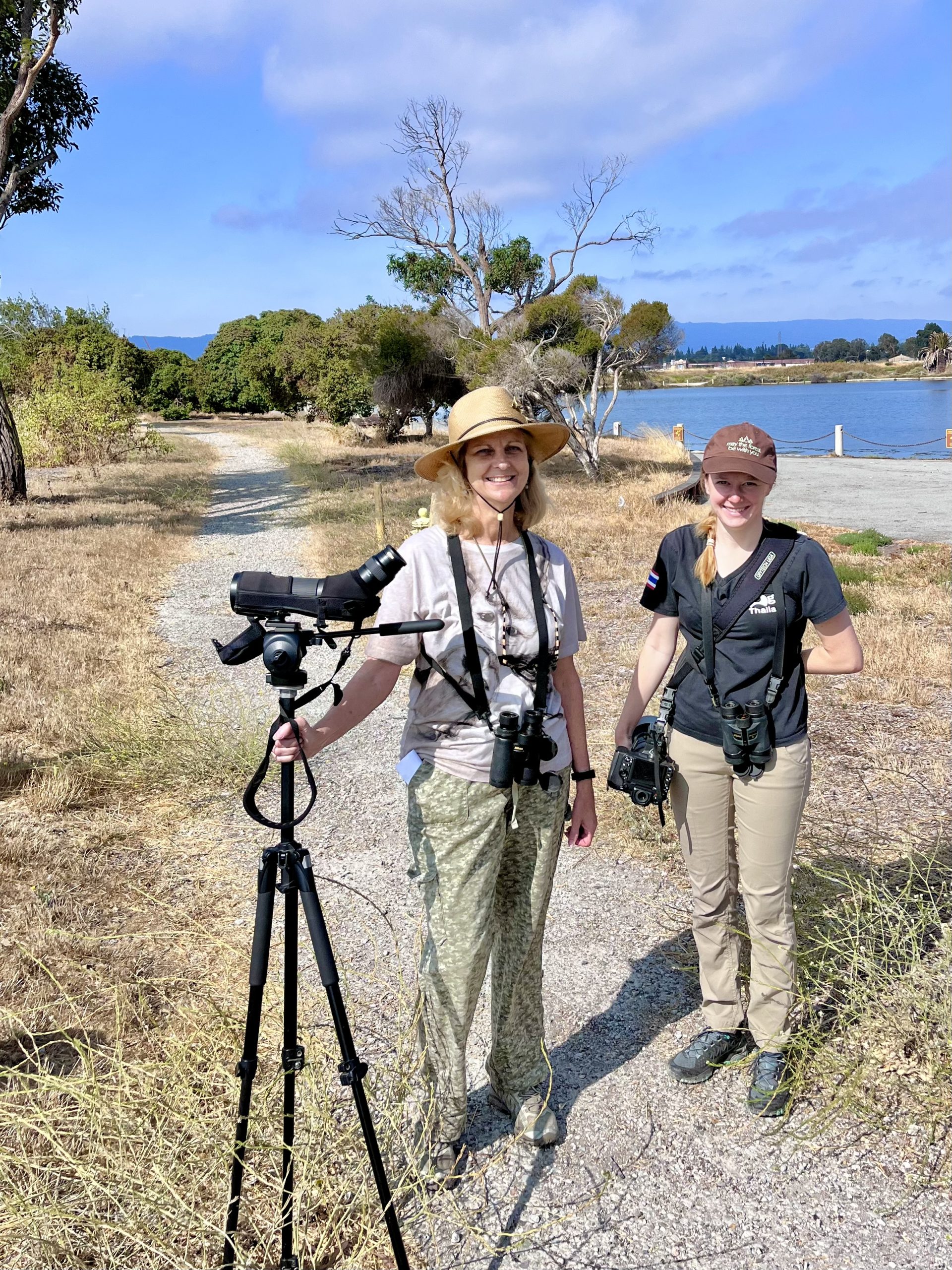 Two women stand on a gravel path with binoculars and tripods, smiling near a lake under a blue sky, wearing outdoor gear and hats.